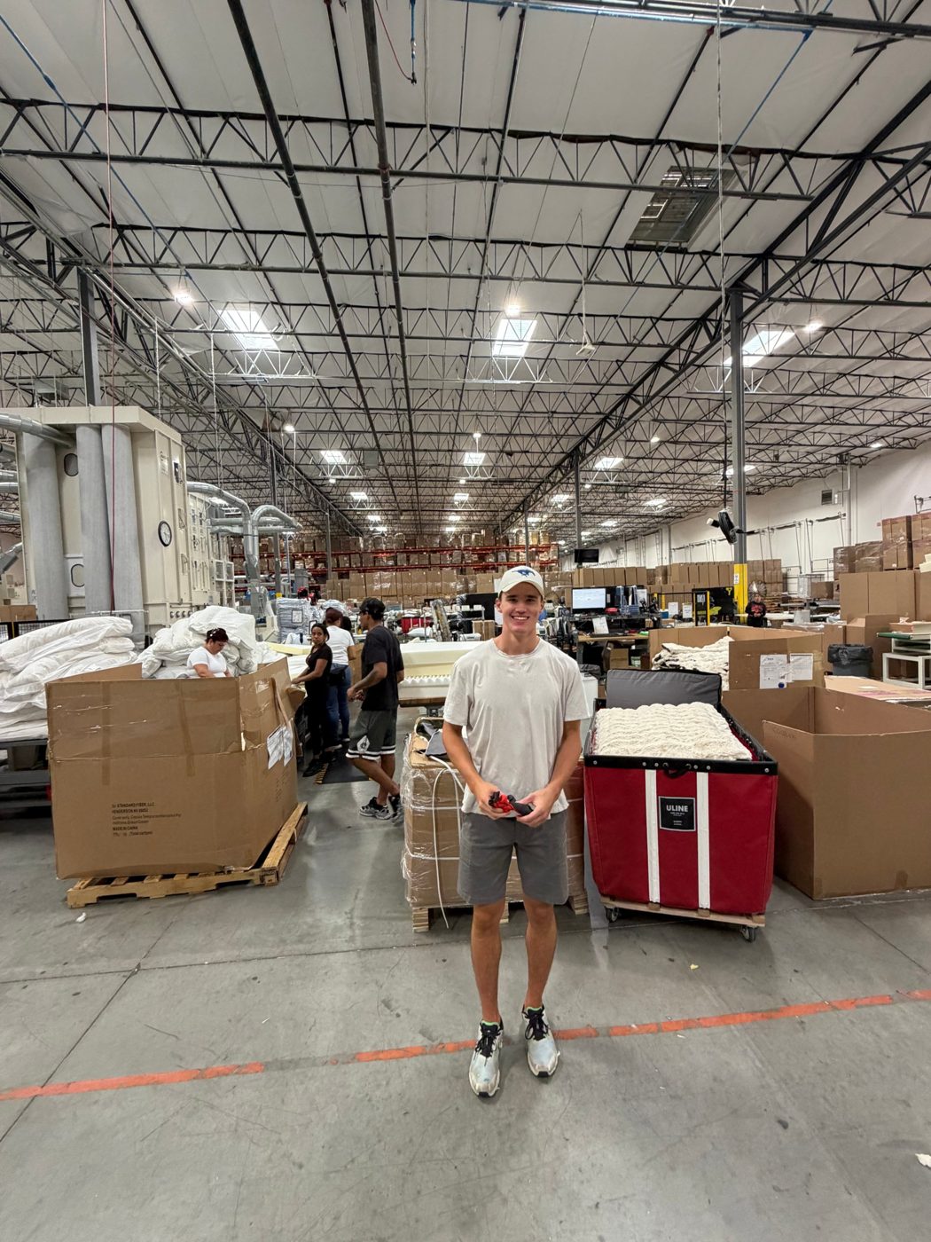 A young man standing on a factory floor among boxes of pillows and bedding