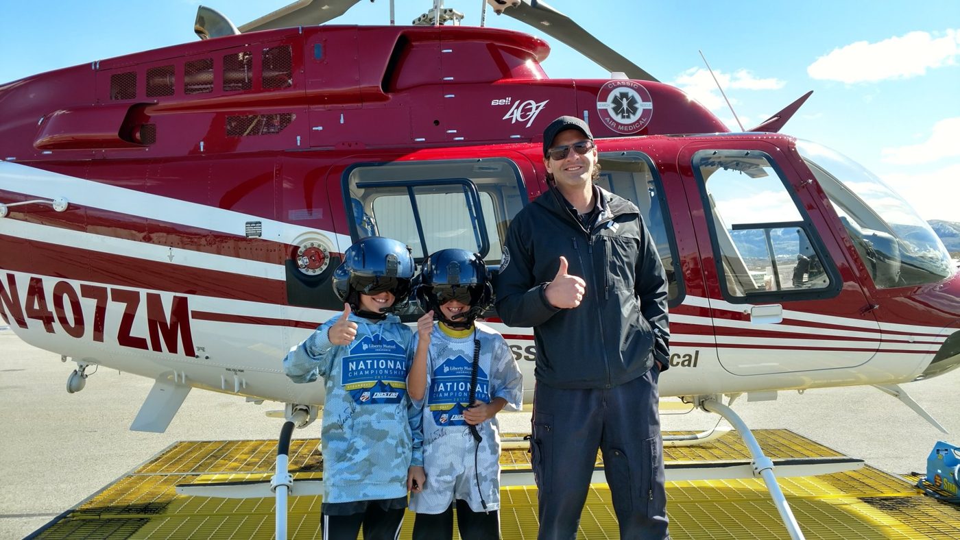Two children in flight helmets giving thumbs up beside a pilot and a red air-medical helicopter