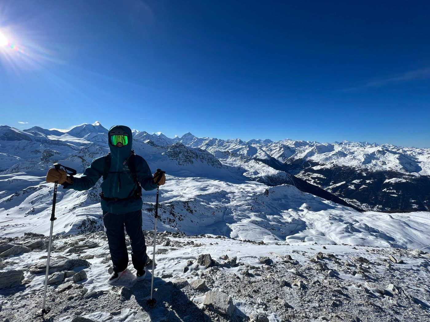 A person in a hooded jacket and goggles standing on a rocky summit above a wide range of snow-covered peaks